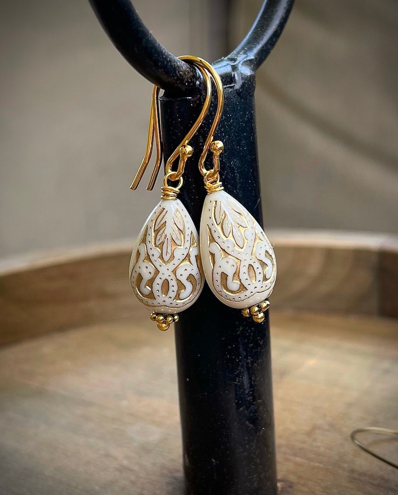 May include: A pair of gold dangle earrings with white teardrop-shaped beads. The beads have a delicate, intricate design etched into them.