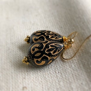 May include: A pair of gold and black teardrop earrings with a delicate filigree design. The earrings are made of metal and have a dangling style.