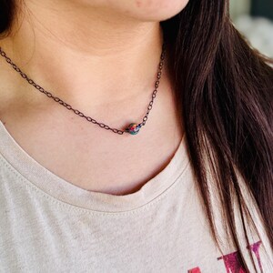 Pu&ograve; includere: Una delicata collana a catena d'argento con un ciondolo a forma di perla iridescente. La perla ha una base verde scuro con sfumature di rosso, arancione e giallo.
