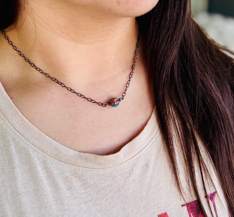 Pu&ograve; includere: Una delicata collana a catena d'argento con un ciondolo a forma di perla iridescente. La perla ha una base verde scuro con sfumature di rosso, arancione e giallo.
