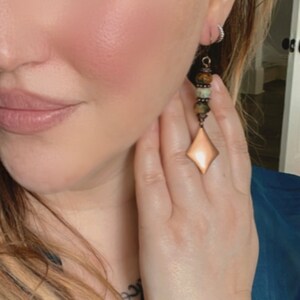 May include: A pair of long, dangle earrings with a copper-toned, diamond-shaped pendant. The earrings feature a mix of green, brown, and copper-toned beads.