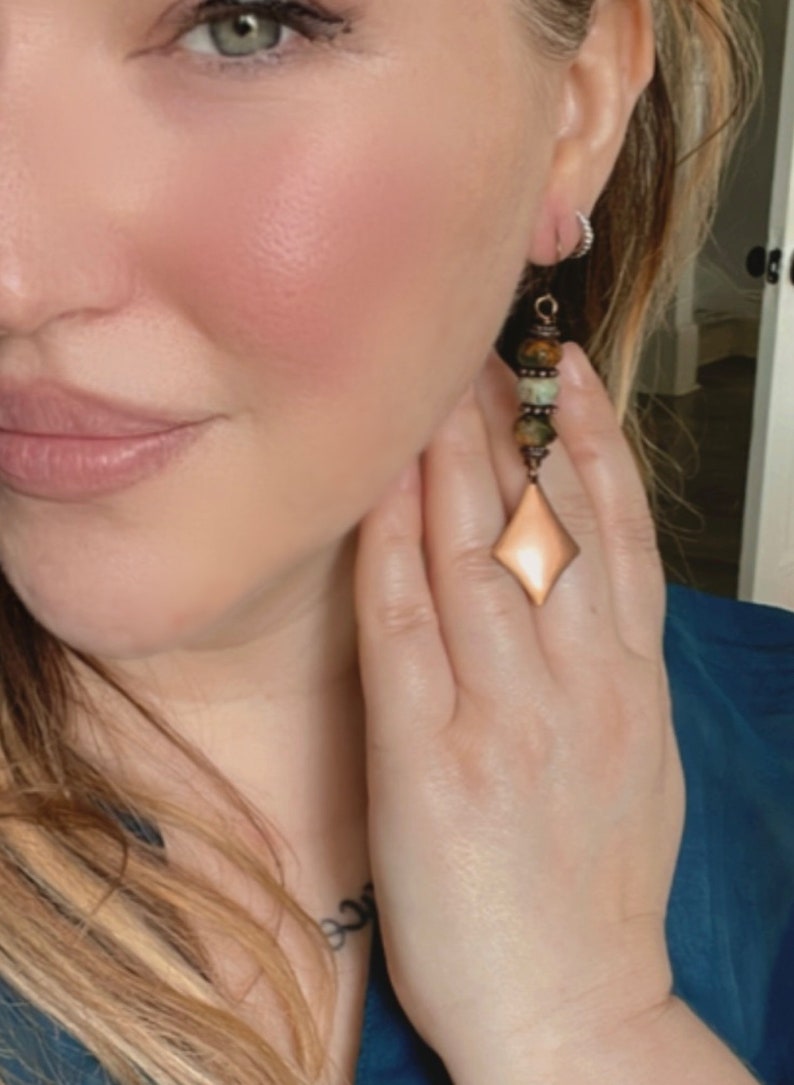 May include: A pair of long, dangle earrings with a copper-toned, diamond-shaped pendant. The earrings feature a mix of green, brown, and copper-toned beads.