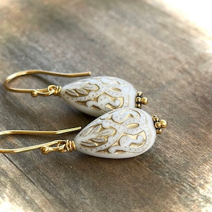 May include: A pair of gold dangle earrings with white teardrop-shaped beads. The beads have a gold filigree design.