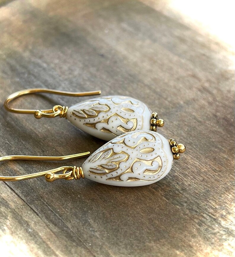 May include: A pair of gold dangle earrings with white teardrop-shaped beads. The beads have a gold filigree design.