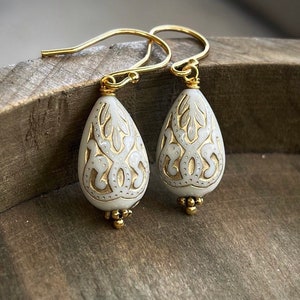 May include: A pair of white teardrop-shaped earrings with gold filigree designs and gold hooks.