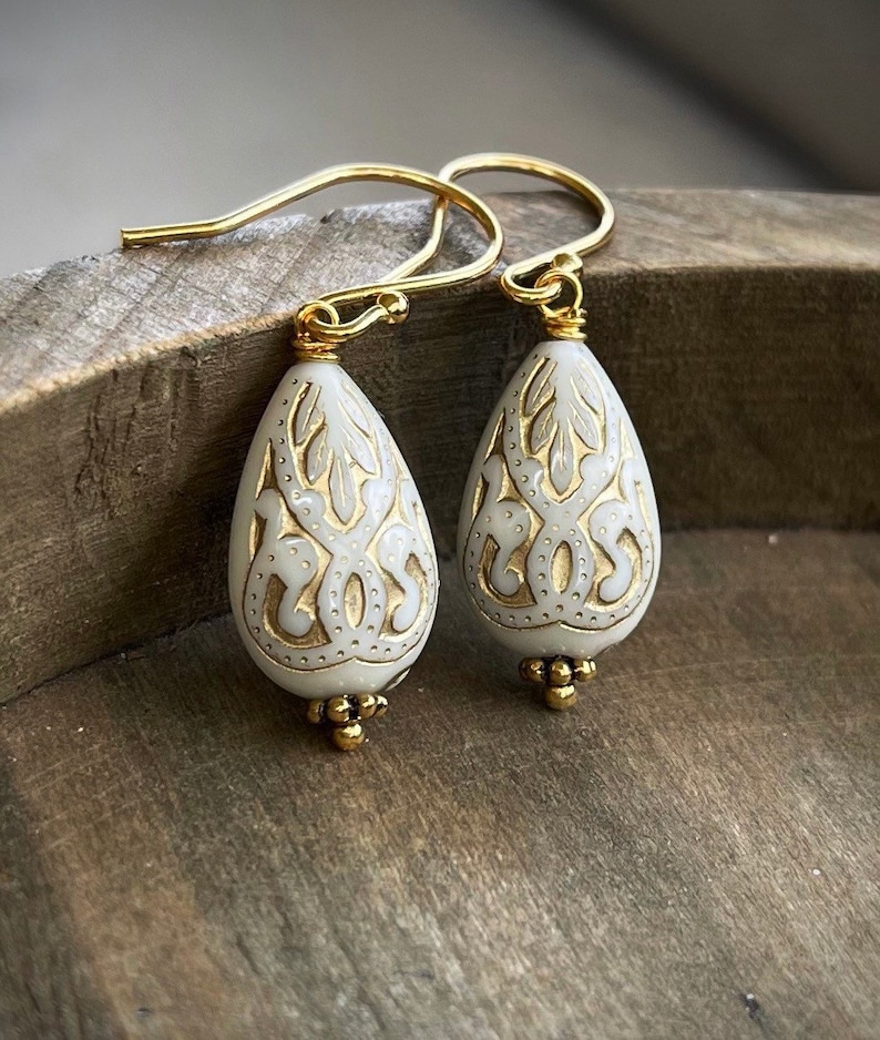 May include: A pair of white teardrop-shaped earrings with gold filigree designs and gold hooks.