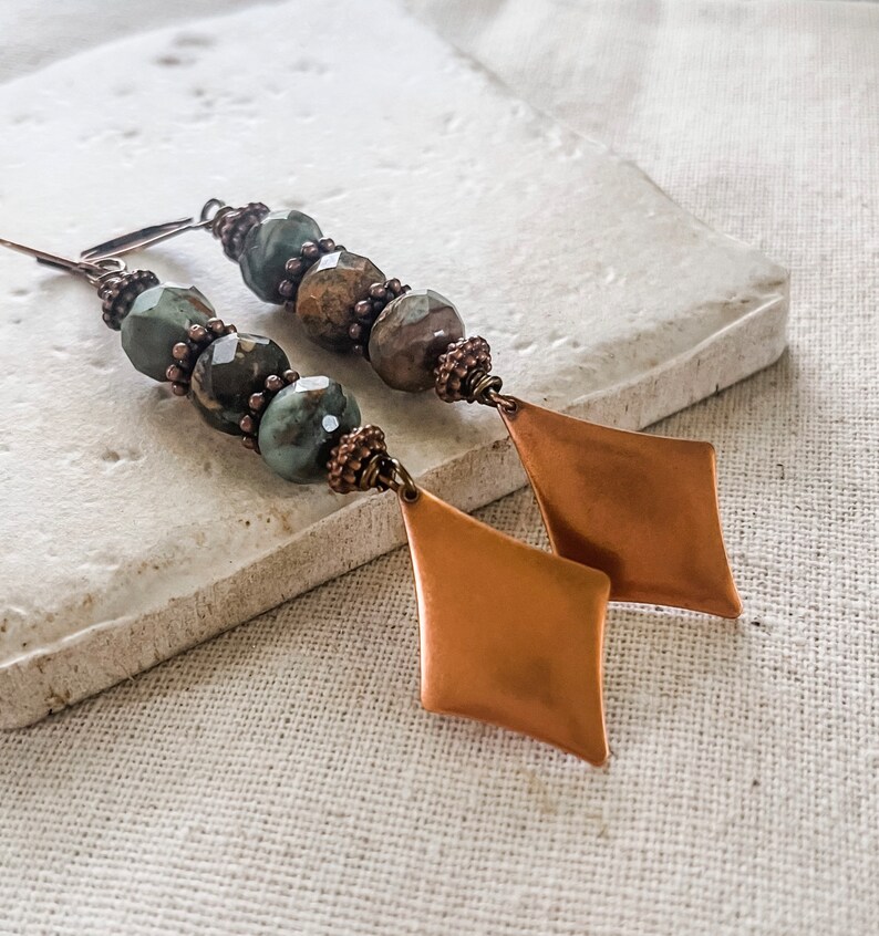 May include: A pair of long, dangle earrings with copper-toned diamond-shaped charms and green and brown gemstone beads.