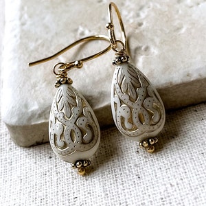 May include: A pair of gold dangle earrings with white teardrop-shaped beads. The beads have intricate gold filigree designs.