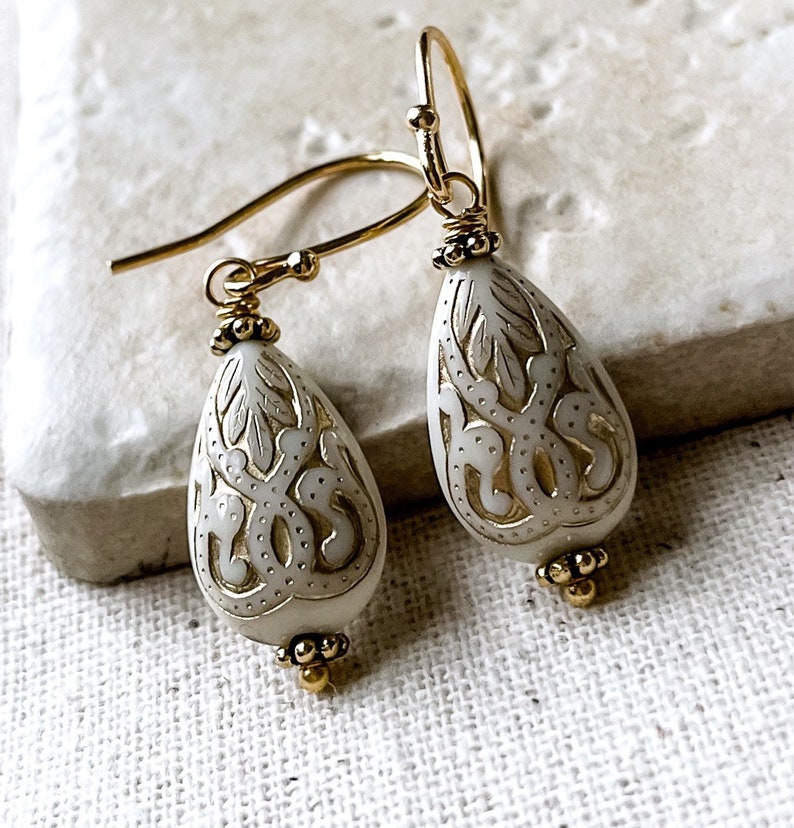 May include: A pair of gold dangle earrings with white teardrop-shaped beads. The beads have intricate gold filigree designs.