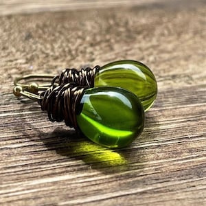Pode incluir: Um par de brincos de gota de vidro verde com envoltório de fio de latão. Os brincos estão sobre uma superfície de madeira.