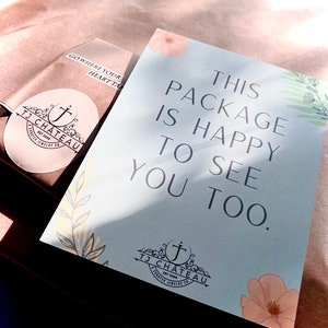 K&ouml;nnte beinhalten: Eine hellblaue Karte mit dem Text "This package is happy to see you too." Die Karte hat ein florales Design mit einem kleinen Logo unten, das "TJ Chateau Est 2008 A Crafted Jewelry Co." lautet.