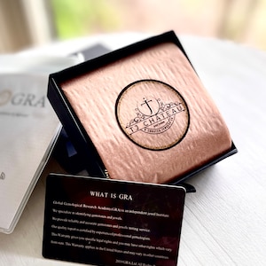 K&ouml;nnte beinhalten: Eine schwarze Box mit braunem Samtbezug und einem goldenen Logo, auf dem "TJ Chateau Est 2009 A Crafted Jewelry Co." steht. Die Box ist ge&ouml;ffnet und eine schwarze Karte mit wei&szlig;em Text ist sichtbar. Die Karte sagt "What is GRA? Global Gemological Research Academy (GRA) is an independent jewel institute. We specialize in identifying gemstones and jewels. We provide reliable and accurate gemstones and jewels testing service. Our quality report is certified by experienced professional gemologists. This Warranty gives you specific legal rights and you may have other rights which vary from state. This Warranty applies in the United States and may vary in other countries. 2019 GRA.Ltd. All Rights Reserved."