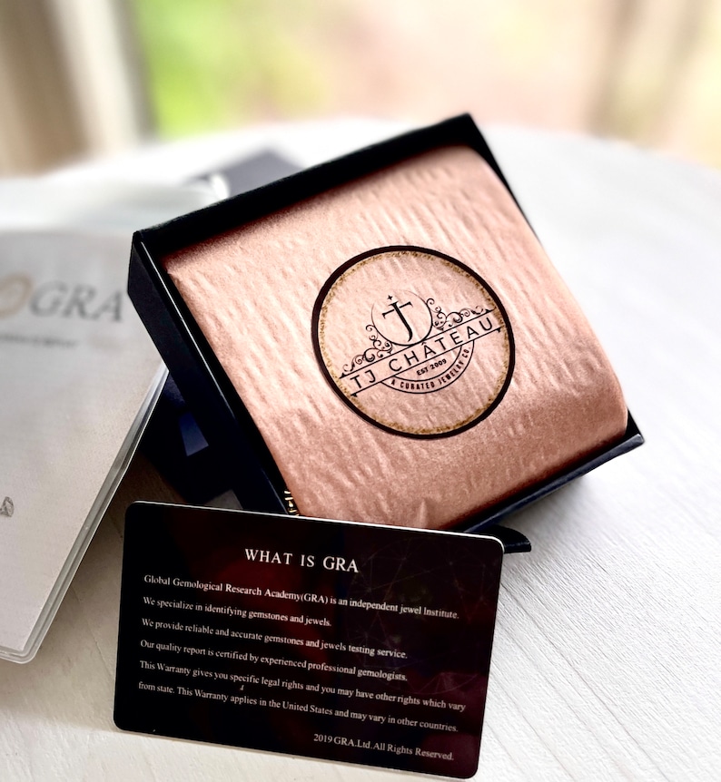 K&ouml;nnte beinhalten: Eine schwarze Box mit braunem Samtbezug und einem goldenen Logo, auf dem "TJ Chateau Est 2009 A Crafted Jewelry Co." steht. Die Box ist ge&ouml;ffnet und eine schwarze Karte mit wei&szlig;em Text ist sichtbar. Die Karte sagt "What is GRA? Global Gemological Research Academy (GRA) is an independent jewel institute. We specialize in identifying gemstones and jewels. We provide reliable and accurate gemstones and jewels testing service. Our quality report is certified by experienced professional gemologists. This Warranty gives you specific legal rights and you may have other rights which vary from state. This Warranty applies in the United States and may vary in other countries. 2019 GRA.Ltd. All Rights Reserved."
