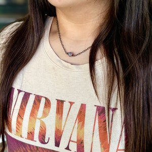 Pu&ograve; includere: Una persona indossa una maglietta beige con la parola "NIRVANA" stampata in arancione, giallo e viola sul davanti. La persona indossa una delicata collana a catena nera con un piccolo ciondolo iridescente.