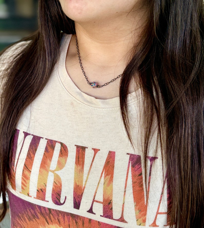 Pu&ograve; includere: Una persona indossa una maglietta beige con la parola "NIRVANA" stampata in arancione, giallo e viola sul davanti. La persona indossa una delicata collana a catena nera con un piccolo ciondolo iridescente.