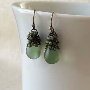 Pode incluir: Um par de brincos pendentes com contas de vidro fosco verde-s&aacute;lvia em forma de gota. O topo de cada conta &eacute; envolto em fio de tom bronze escuro e acentuado com pequenas contas verde-s&aacute;lvia correspondentes, suspensas em ganchos de bronze.