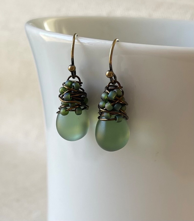Pode incluir: Um par de brincos pendentes com contas de vidro fosco verde-s&aacute;lvia em forma de gota. O topo de cada conta &eacute; envolto em fio de tom bronze escuro e acentuado com pequenas contas verde-s&aacute;lvia correspondentes, suspensas em ganchos de bronze.