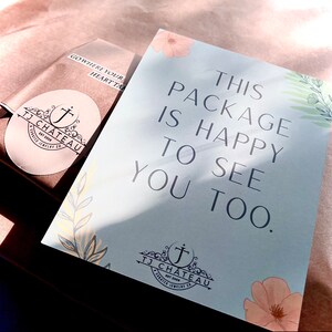 Pu&ograve; includere: Un biglietto azzurro chiaro con il testo "This package is happy to see you too." Il biglietto ha un design floreale con un piccolo logo in basso a destra che dice "TJ Chateau Est 2009 A curated jewelry co."