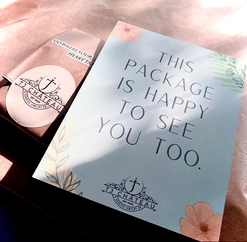 Pu&ograve; includere: Un biglietto azzurro chiaro con il testo "This package is happy to see you too." Il biglietto ha un design floreale con un piccolo logo in basso a destra che dice "TJ Chateau Est 2009 A curated jewelry co."
