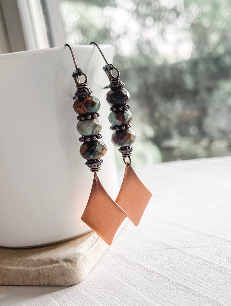 May include: A pair of dangle earrings with copper-toned diamond-shaped charms and brown, green, and blue gemstone beads.