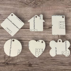 May include: A collection of blank, cream-colored tags in various shapes: a square, a house, a rectangle, a circle, a heart, and a dog bone. Each tag has a hole for hanging and the text "protective film, please tear off".