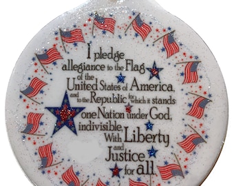 May include: A white, round ornament with the text of the Pledge of Allegiance. The ornament is decorated with small American flags and stars in red, white, and blue. A gold string is attached for hanging.