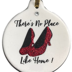 May include: White, round ornament with the text "There's No Place Like Home!" and a pair of red glitter high-heeled shoes. The ornament has a gold ribbon for hanging and a small rhinestone.