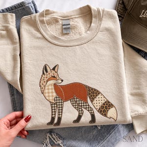 May include: A sand-colored crewneck sweatshirt featuring a central graphic of a fox. The fox illustration is composed of various patterned sections including plaid, houndstooth, leopard print, and solid rust-orange fabric textures.