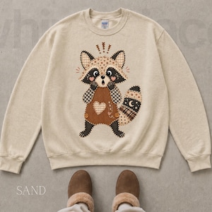 May include: A sand-colored crewneck sweatshirt featuring a central graphic of a surprised raccoon. The raccoon is depicted with a patchwork-style design in shades of brown, cream, and black, wearing overalls with a heart and small buttons.