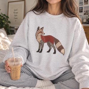 Puede incluir: Una sudadera gris claro de cuello redondo con un gráfico central de un zorro. La ilustración del zorro está compuesta por varias secciones estampadas que incluyen estampado de leopardo, cuadros y segmentos de color óxido en el cuerpo.