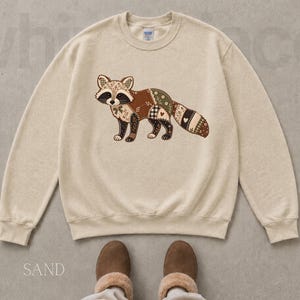 May include: A sand-colored crewneck sweatshirt featuring a graphic of a raccoon designed with a patchwork pattern of brown, green, and cream floral and checkered fabrics. The raccoon has a striped tail and a small bow at its neck.