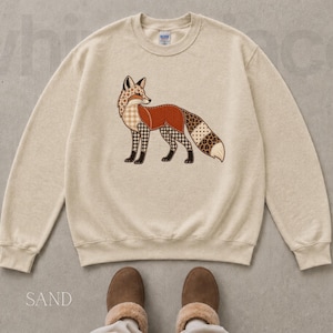 May include: A sand-colored crewneck sweatshirt featuring a central graphic of a fox. The fox is composed of various patterned sections, including plaid, leopard print, and solid rust-orange segments, with a white-tipped tail and black outlines.