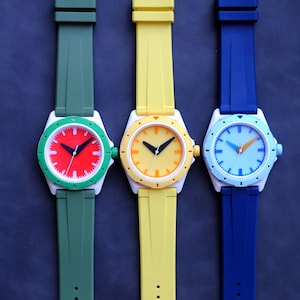 May include: Three wristwatches with white cases and colorful faces. The watches have green, yellow, and blue bands.