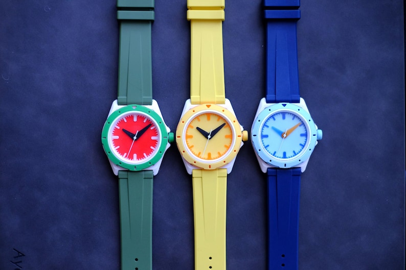 May include: Three wristwatches with white cases and colorful faces. The watches have green, yellow, and blue bands.