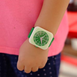 Op de afbeelding: Een groen-witte polshorloge met een uniek wijzerplaatontwerp. Het horloge heeft een groene band en een witte wijzerplaat met zwarte cijfers en wijzers.