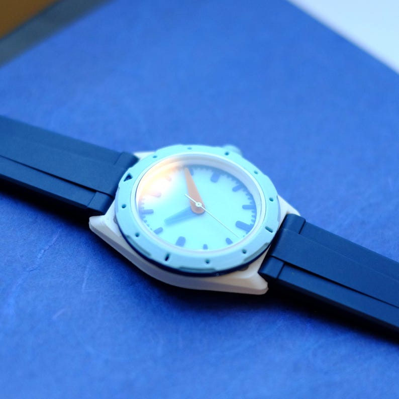 May include: A white and blue wristwatch with a blue face and orange hands. The watch has a black rubber strap.