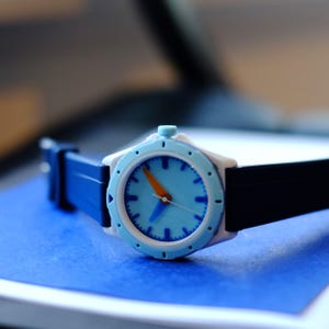 May include: A blue and white wristwatch with an orange second hand and a black strap. The watch face has a light blue background with white markings.