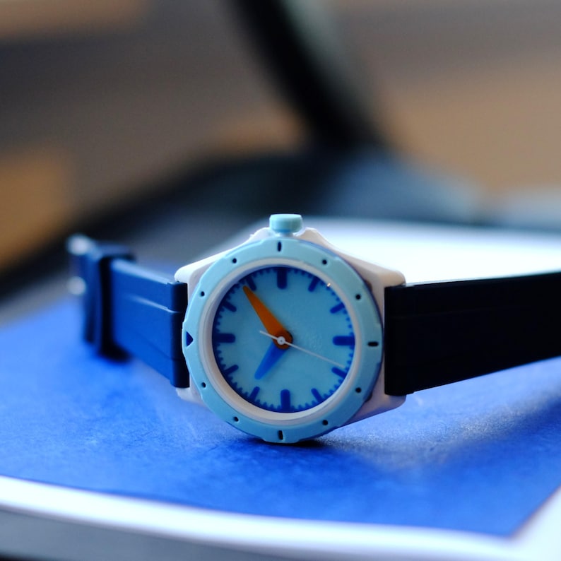 May include: A blue and white wristwatch with an orange second hand and a black strap. The watch face has a light blue background with white markings.