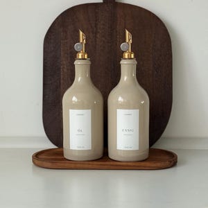 Könnte beinhalten: Zwei beigefarbene Keramikflaschen mit goldenen Ausgüssen und weißen Etiketten, eine mit der Aufschrift "OL" und die andere mit "ESSIG". Jede Flasche fasst 500 ml. Sie stehen auf einem Holztablett vor einem dunklen Holzschneidebrett.