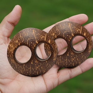 May include: Two round, dark brown coconut shell pieces with two circular holes each. The natural wood grain is visible, with lighter brown and tan patterns. The pieces are held in a hand against a blurred green background.