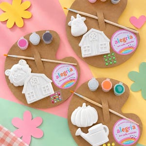May include: Three brown cardboard palettes, each featuring a white paintable figurine, a wooden paintbrush, three small paint pots, and a circular sticker with the text "alegria em cada caixinha". The kits include small adhesive gems for decoration.