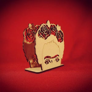 May include: Wooden napkin holder featuring a cutout design of Frida Kahlo with a flower crown. The holder is made of natural wood and has a brown finish.