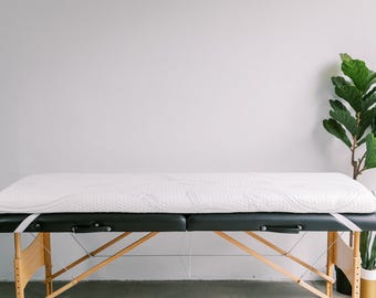 May include: A white padded massage table with a black frame and wooden legs. The table is set up in a room with a white wall and a potted plant in the background.