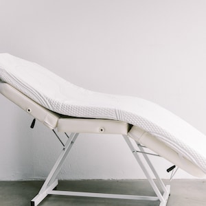 May include: White adjustable massage table with a padded, quilted top. The table is in a raised position and has a white metal frame.