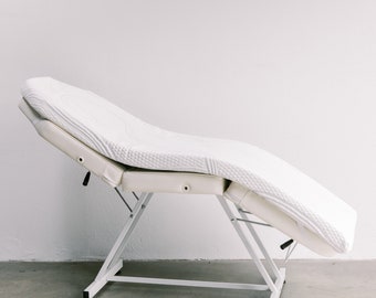May include: White adjustable massage table with a padded, quilted top. The table is in a raised position and has a white metal frame.