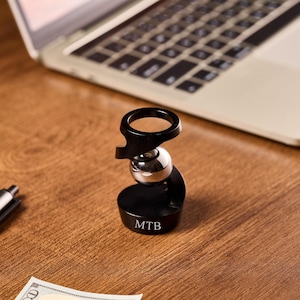 May include: A black and silver desk accessory with the initials "MTB" engraved on the base. The accessory has a ring-shaped top, a silver ball in the middle, and a curved base. A laptop and pen are in the background.