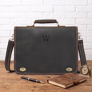 May include: A dark brown leather briefcase featuring a top handle, a detachable shoulder strap, and two brass-toned metal turn-lock closures. The front is engraved with the letter W and the name William. A pen and a notebook are nearby.