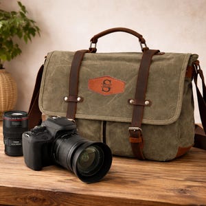 Puede incluir: Bolsa de cámara de lona verde oliva con detalles en cuero marrón, correas y asa. Cuenta con un parche de cuero personalizado con iniciales. Una cámara DSLR negra y una lente están junto a la bolsa, lo que sugiere equipo de fotografía.