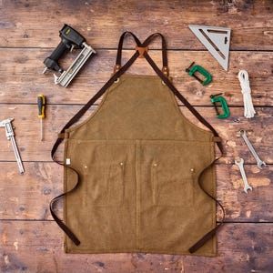 May include: A brown canvas apron with leather straps and multiple pockets, surrounded by tools including a nail gun, wrenches, and a speed square. The apron is designed for woodworking or other craft projects.