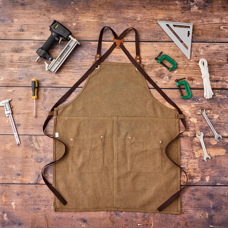 May include: A brown canvas apron with leather straps and multiple pockets, surrounded by tools including a nail gun, wrenches, and a speed square. The apron is designed for woodworking or other craft projects.